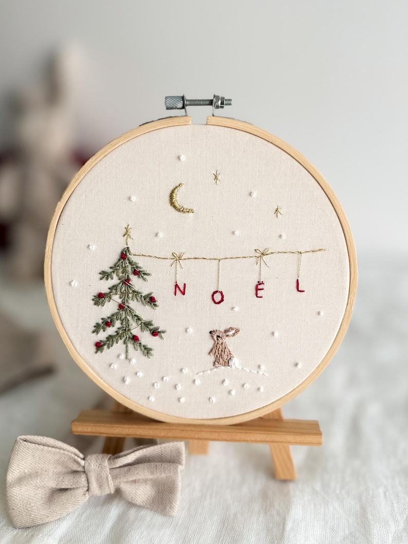 Może przedstawiać: Haftowany obręcz z zielonym drzewem bożonarodzeniowym, białym kr&oacute;likiem i napisem "NOEL" czerwonymi literami. Obręcz znajduje się na drewnianym sztalugach.