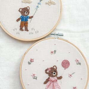 May include: Two embroidered hoops featuring teddy bears. One bear holds a kite, the other a balloon. The kite is blue and green, the balloon is pink. The bears are brown, wearing blue and orange, and pink dresses. The background is white.