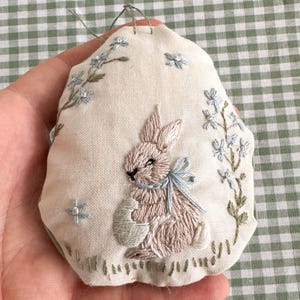 Puede incluir: Un huevo de tela cosido a mano con un conejito marrón claro que sostiene un huevo blanco. El conejito está bordado con hilo rosa y azul claro. El huevo está decorado con flores azules y verdes.