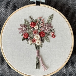 Puede incluir: Un aro de bordado hecho a mano con un ramo de flores con flores rosas, rojas y blancas, hojas verdes y una cinta rosa.