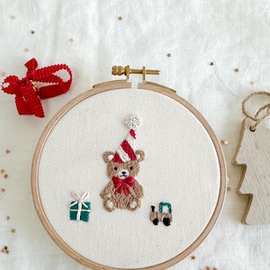 Peut inclure: Art de broderie sur un cercle en bois avec un ourson brun portant un chapeau rayé rouge et blanc, tenant un nœud rouge. L'ourson est entouré d'une boîte cadeau verte et d'un camion jouet vert. Le cercle est en bois et a un fond en tissu blanc.