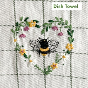 Peut inclure: Une serviette de cuisine blanche avec un motif vichy vert. Un motif en forme de c&oelig;ur est brod&eacute; sur la serviette avec une abeille au centre et des fleurs autour de l'abeille. Le texte "Dish Towel" est brod&eacute; en lettres vertes dans le coin sup&eacute;rieur droit.