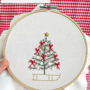 Op de afbeelding: Een borduurring met een kerstboomontwerp. De boom is groen met rode strikken en witte ornamenten. Een gouden ster bekroont de boom en een gouden slee staat aan de basis. De tekst "Step-by-Step Instructions" en "PDF Pattern" is ook zichtbaar.