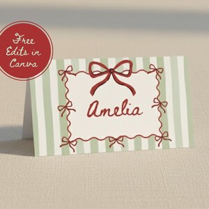 May include: A folded place card with a green and white striped background. The card features a red bow and the name "Amelia" in a handwritten font. A red circle with the text "Free Edits in Canva" is in the upper left corner.