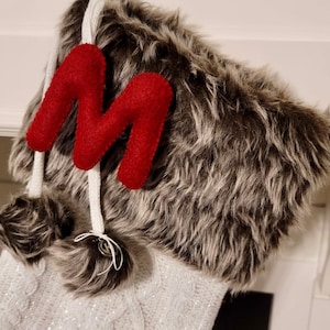 May include: White and gray faux fur stocking with a red felt letter M on the front. The stocking has a white pom-pom on the top.