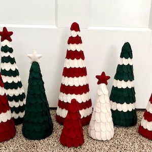 May include: A collection of decorative felt Christmas trees in various sizes and colors. The trees are primarily red, white, and green, with some topped with star ornaments. The trees are arranged on a neutral-colored surface.