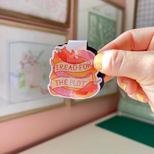 May include: A colorful sticker with the text "I READ FOR THE PLOT" on a banner, featuring red chili peppers and flames. The sticker is held up against a background with framed artwork.