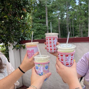 May include: Four friends raise their iced coffee drinks in a toast. Each drink has a different patterned reusable cup sleeve. One sleeve has a pink and white cherry pattern, another has a pink and white seashell pattern, and another has a pink and white "Drink Local Coffee" pattern.
