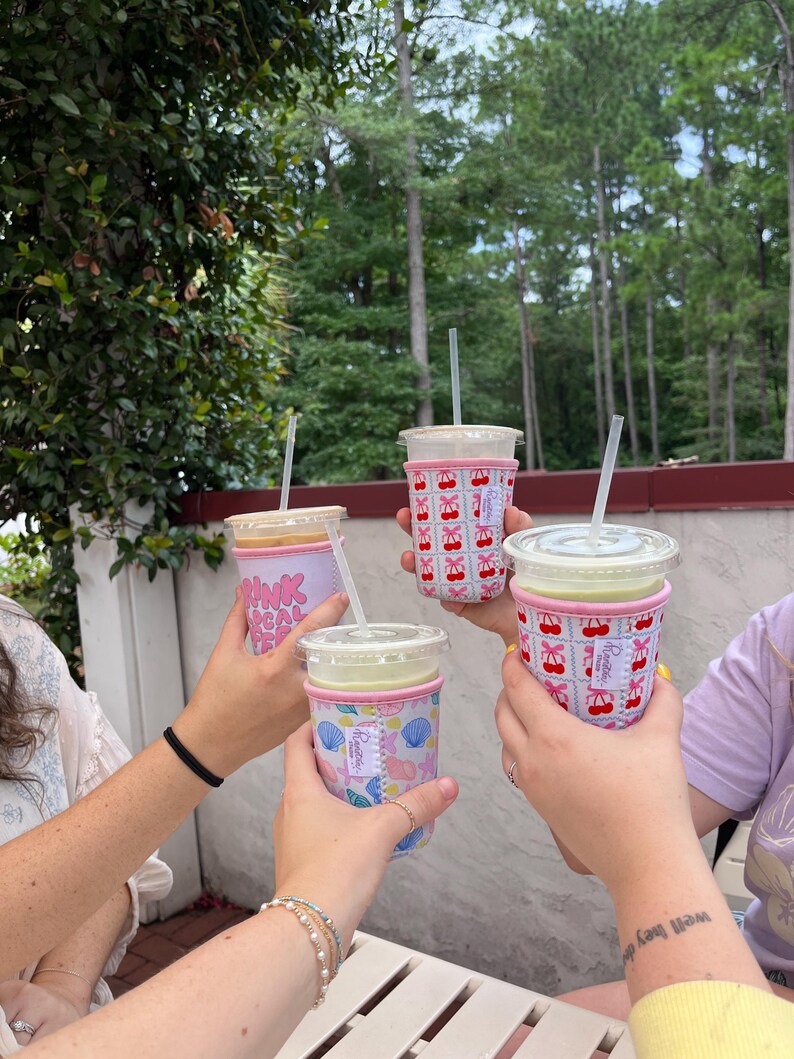 May include: Four friends raise their iced coffee drinks in a toast. Each drink has a different patterned reusable cup sleeve. One sleeve has a pink and white cherry pattern, another has a pink and white seashell pattern, and another has a pink and white "Drink Local Coffee" pattern.