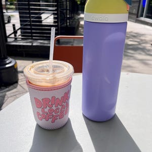 May include: A purple reusable water bottle with a yellow lid and a white label that says "owala". The bottle is sitting on a white table next to a pink and white insulated cup with the text "Drink Local Coffee".