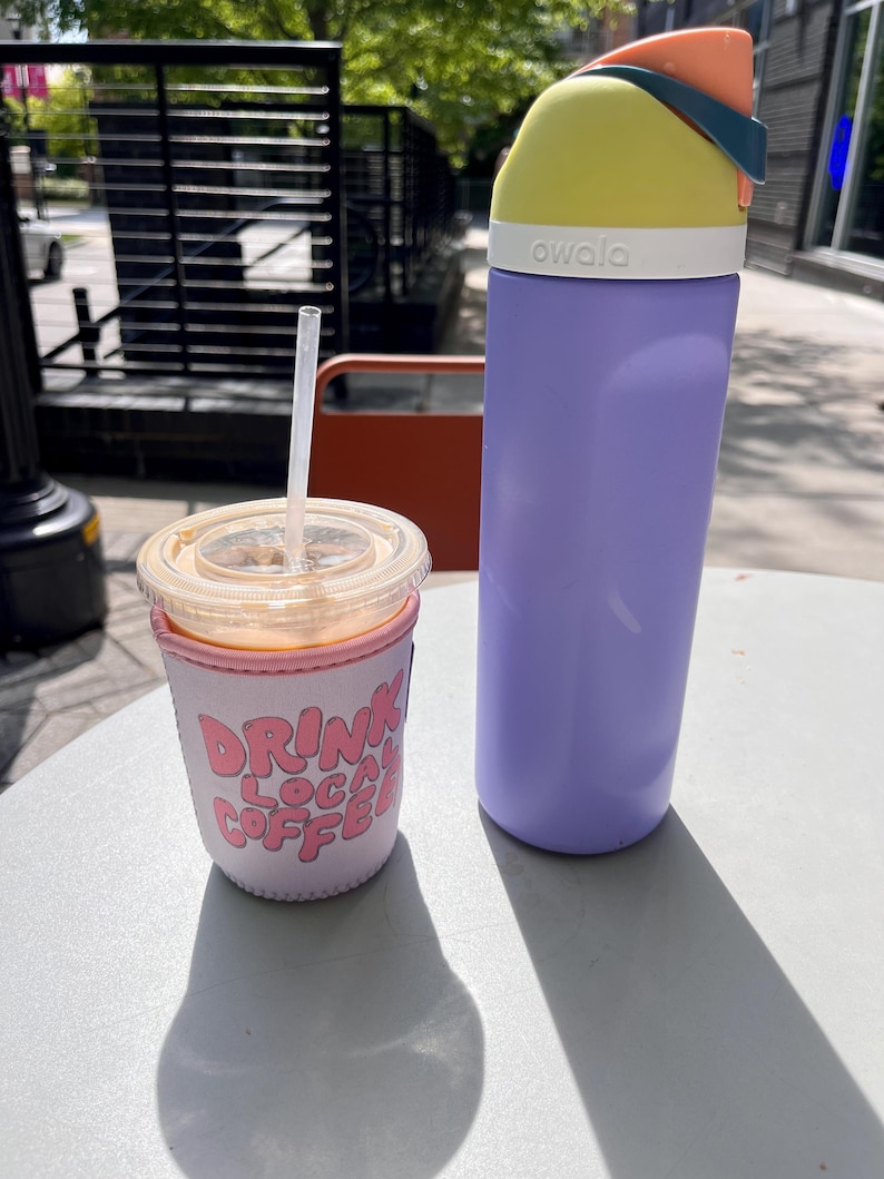 May include: A purple reusable water bottle with a yellow lid and a white label that says "owala". The bottle is sitting on a white table next to a pink and white insulated cup with the text "Drink Local Coffee".