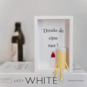 Könnte beinhalten: Ein weißer Rahmen mit dem Text "Drinks de eijne met?" und einer roten Grafik. Eine gelbe Figur, die einem Bierglas ähnelt, sitzt auf dem Rahmen. Das Schild liegt auf einem Stapel weißer Bücher.