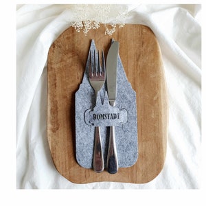 Könnte beinhalten: Ein grauer Filz-Besteckhalter in Form eines Hauses mit dem aufgedruckten Wort "Domstadt". Eine Gabel und ein Messer sind im Halter platziert.