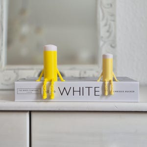 Könnte beinhalten: Zwei gelb-weiße Bier-Buchstützen mit Armen und Beinen sitzen auf einem weißen Buch. Auf dem Buch steht das Wort "WHITE" auf dem Einband. Diese Buchstützen sind eine lustige und skurrile Ergänzung für jedes Bücherregal.