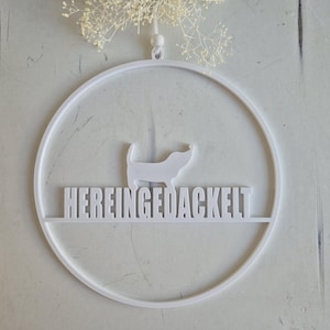 Könnte beinhalten: Weißer, kreisförmiger Wandschmuck mit einer Dackel-Silhouette und dem deutschen Schriftzug "HEREINGEDACKELT". Das Design wird von einer kleinen Schlaufe gehalten und mit Trockenblumen verziert.