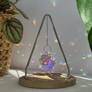 Op de afbeelding: Een heldere kristallen bol hangt aan een zilveren metalen frame met een houten basis. De kristallen bol is facetgeslepen en reflecteert regenbooglichtpatronen. Een kleine plant in een geweven mand staat op de achtergrond.
