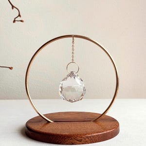 May include: A clear crystal ball hangs from a gold-colored metal ring and chain, suspended within a larger gold-colored metal hoop. The hoop is mounted on a round, dark wooden base. The background is a soft, neutral color.
