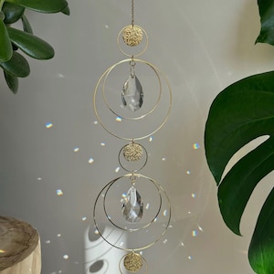 Op de afbeelding: Een decoratieve hangende kristallen zonnevanger met goudkleurige metalen ringen en getextureerde cirkelvormige accenten. De zonnevanger heeft twee heldere, druppelvormige kristallen en hangt aan een ketting. De achtergrond bevat groene planten en lichtreflecties.