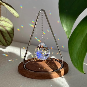 Può includere: Una sfera di cristallo trasparente è sospesa a un telaio in metallo argentato, appoggiata su una base rotonda in legno. Il cristallo rifrange la luce, creando motivi arcobaleno. Un oggetto decorativo per la casa.