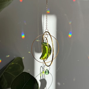 Op de afbeelding: Een hangende zonnevanger met een goudkleurige ketting en een cirkelvormig frame. Een groene, halvemaanvormige kristal hangt in het frame, met een kleinere groene kristal eronder. De zonnevanger is ontworpen om zonlicht op te vangen en regenbogen te creëren.
