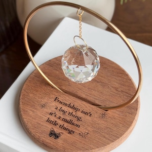 May include: A clear crystal ball suspended from a gold-coloured metal hoop, set on a round wooden base. The base is engraved with the quote, "Friendship isn't a big thing, it's a million little things," and butterfly and star motifs.