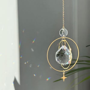 Op de afbeelding: Een heldere kristallen zonnevanger met een facetgeslepen, bolvormig kristal dat is opgehangen in goudkleurige ringen en een sterbedel. De zonnevanger hangt aan een fijne gouden ketting en reflecteert licht, waardoor regenbogen ontstaan.