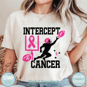 以下が含まれることがあります： クリーム色のTシャツ。デザインは、黒で「INTERCEPT CANCER」、ピンクのフットボール、フットボール選手のシルエット、ピンクのリボンが特徴です。シャツには「SVG」と「Digital Download」の文字も入っています。