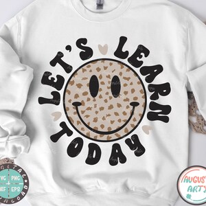 Puede incluir: Sudadera blanca con un diseño gráfico en negro y marrón. El gráfico presenta una cara sonriente con un patrón de estampado de leopardo y el texto "Let's Learn Today" en una fuente retro.