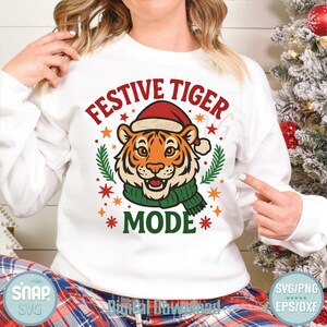 May include: White sweatshirt with a graphic of a tiger wearing a Santa hat and green scarf. The text reads "Festive Tiger Mode" with star and leaf accents. A fun, holiday-themed design.