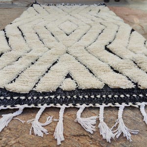 May include: A rectangular rug with a geometric design. The rug features a cream-coloured, textured pattern against a black background. White tassels adorn the bottom edge. The rug is made of a soft, woven material.
