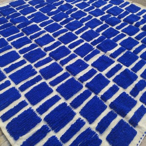 Blue and white Moroccan hand-knotted wool rug with a geometric grid of blue blocks on white, placed on tiled floor.