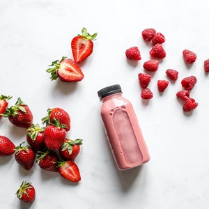 Może przedstawiać: R&oacute;żowy koktajl w przezroczystym plastikowym butelce z czarną zakrętką, otoczony świeżymi truskawkami i malinami na białej marmurowej powierzchni. Truskawki są całe i pokrojone, a maliny są rozrzucone.
