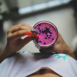 Może przedstawiać: Szklanka żywego fioletowego smoothie, udekorowana świeżymi jagodami, trzymana przez osobę. Smoothie ma bogatą, kremową konsystencję, a do napoju włożona jest czerwona słomka. Osoba ma na sobie białą koszulkę.