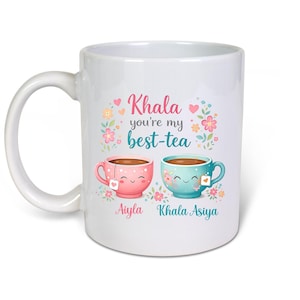 May include: A white ceramic mug featuring a graphic of two smiling tea cups, one pink and one teal, surrounded by small flowers and hearts. The text reads "Khala you're my best-tea" with the names "Aiyla" and "Khala Asiya" printed below the cups.