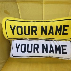 May include: Two rectangular throw cushions with rounded corners, designed to resemble street signs. The top cushion is yellow with black text reading YOUR NAME, and the bottom cushion is white with black text reading YOUR NAME, both outlined in black.