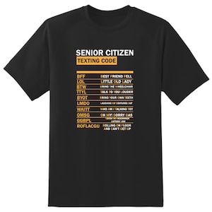 May include: Black t-shirt with a white graphic of a senior citizen texting code. The code includes abbreviations like BFF, LOL, BTW, TTYL, BYOT, LMDO, WAITT, OMSG, GGBPL, and ROFLACGU, with their corresponding meanings.