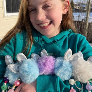 May include: A girl is holding four fluffy pom-pom keychains with silver bunny ears. The keychains are blue, purple, white, and light blue. Each keychain has a beaded charm with colourful beads.