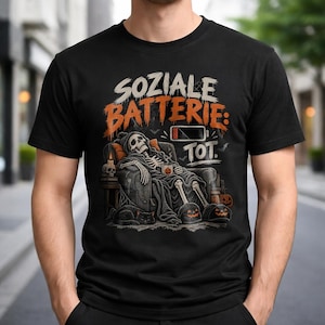 Può includere: Una t-shirt nera con la grafica di uno scheletro che si rilassa su una poltrona con una coperta, una tazza e un gatto. Il testo recita SOZIALE BATTERIE: TOT. con un'icona di batteria scarica, zucche, una candela e dei pipistrelli.