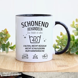 May include: White ceramic mug with a black rim and handle. The mug has black text that reads "Schonend Behandeln Das Unikat ist schon 40 Falten, Nicht Bugeln Nicht Schleudern Nur Handwasche". The mug also has four black and white laundry care symbols.