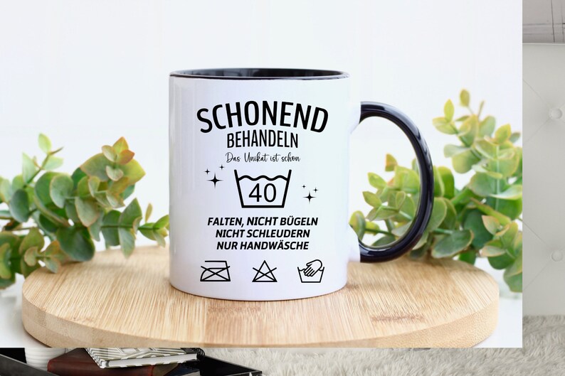 May include: White ceramic mug with a black rim and handle. The mug has black text that reads "Schonend Behandeln Das Unikat ist schon 40 Falten, Nicht Bugeln Nicht Schleudern Nur Handwasche". The mug also has four black and white laundry care symbols.