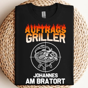 Könnte beinhalten: Schwarzes T-Shirt mit einem Grafikdruck eines Grills mit Flammen und dem Text "Auftrags Griller Johannes am Bratort".