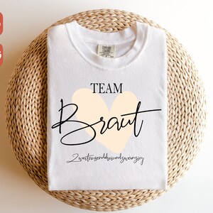 May include: White t-shirt with the text "TEAM Braut" in black lettering with a peach heart. The text "zweittausendundzwanzig" is written in black below the heart.