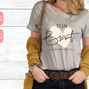 May include: A gray t-shirt with the text "TEAM BRAUT" in black lettering with a pink heart behind the word "BRAUT". The text "Zweitens sind wir nur ganz" is written in black below the heart.