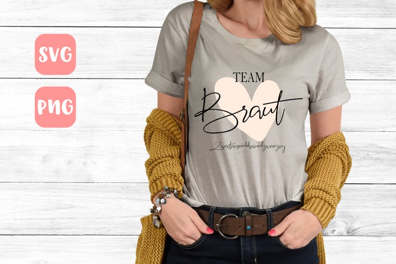 May include: A gray t-shirt with the text "TEAM BRAUT" in black lettering with a pink heart behind the word "BRAUT". The text "Zweitens sind wir nur ganz" is written in black below the heart.