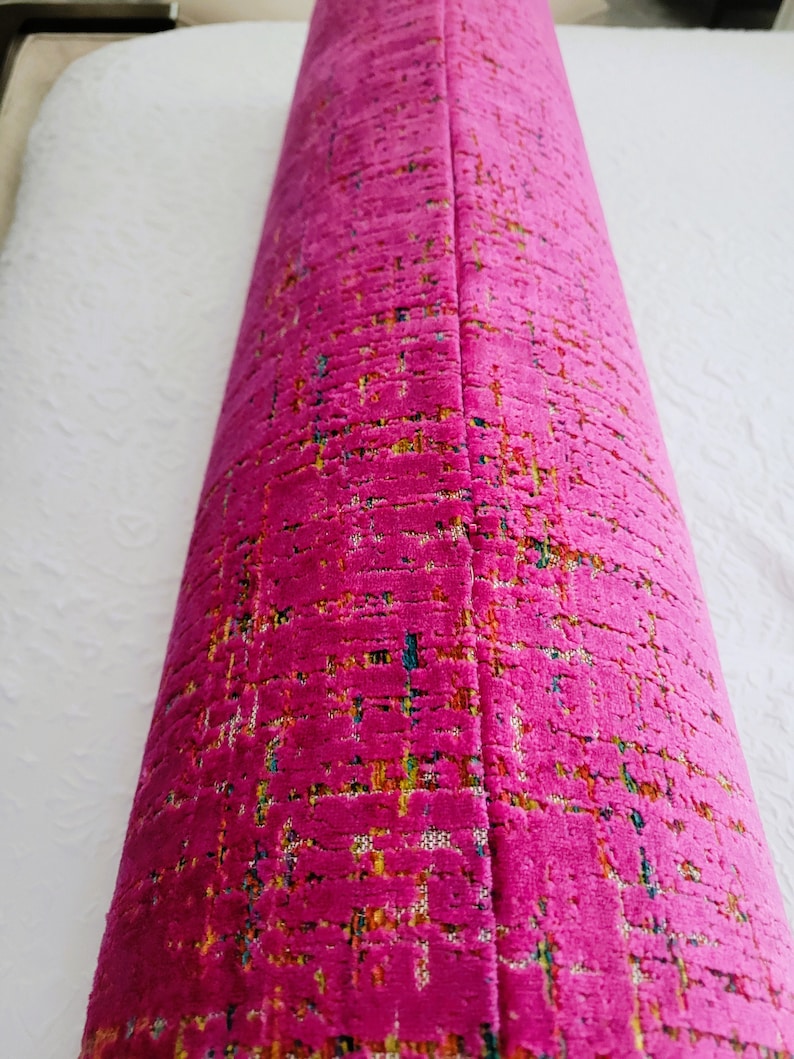 May include: A rolled-up, vibrant fuchsia-colored fabric with a textured, woven appearance. The fabric has a subtle pattern with flecks of yellow, blue, and green. The item appears to be a decorative textile, possibly for upholstery or home decor.