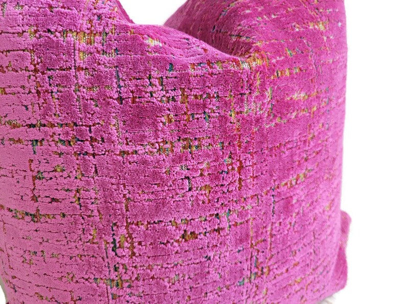 May include: Close-up of a vibrant fuchsia pillow with a textured, woven appearance. The fabric has a grid-like pattern with subtle flecks of yellow, green, and blue, creating a rich, multi-tonal effect. The pillow is a decorative home accent.