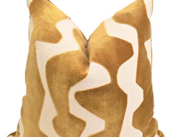 Puede incluir: Un cojín decorativo con un diseño abstracto en dorado y crema. El cojín tiene un aspecto suave y texturizado y una paleta de colores neutros, adecuado para varios estilos de diseño de interiores. El cojín es cuadrado.