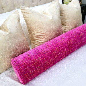 May include: A bright pink bolster pillow with a textured pattern rests on a white bed, alongside three gold-toned square pillows. The bed has a neutral-colored headboard. The bolster pillow adds a pop of color to the neutral bedding.