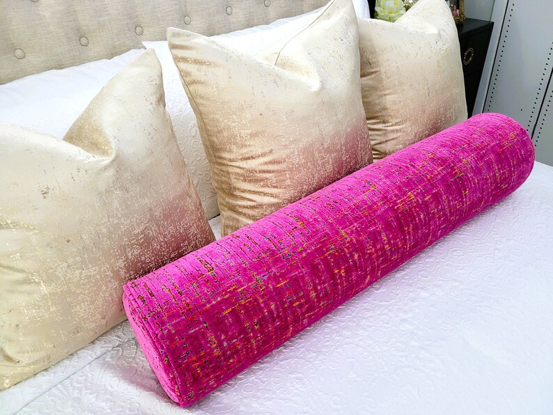 May include: A bright pink bolster pillow with a textured pattern rests on a white bed, alongside three gold-toned square pillows. The bed has a neutral-colored headboard. The bolster pillow adds a pop of color to the neutral bedding.