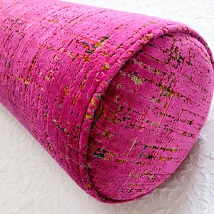 May include: A vibrant magenta bolster pillow with a textured, patterned fabric. The cylindrical pillow features rounded ends and a decorative trim. The fabric has flecks of yellow, green, and blue.